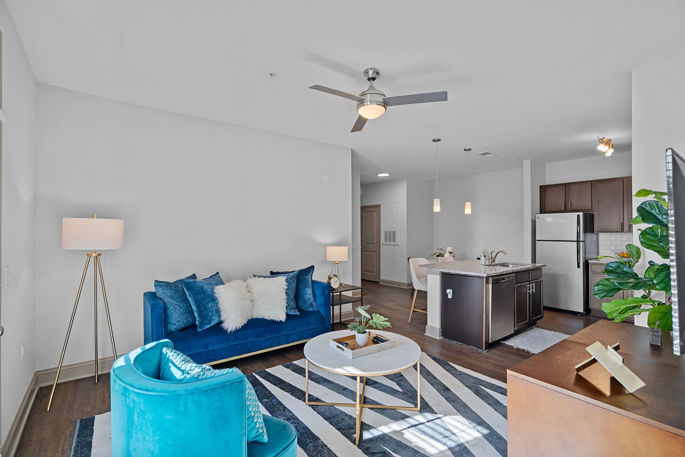 Modern open living room and kitchen with blue velvet seating, dark wood flooring, ceiling fan, and stainless steel appliances.