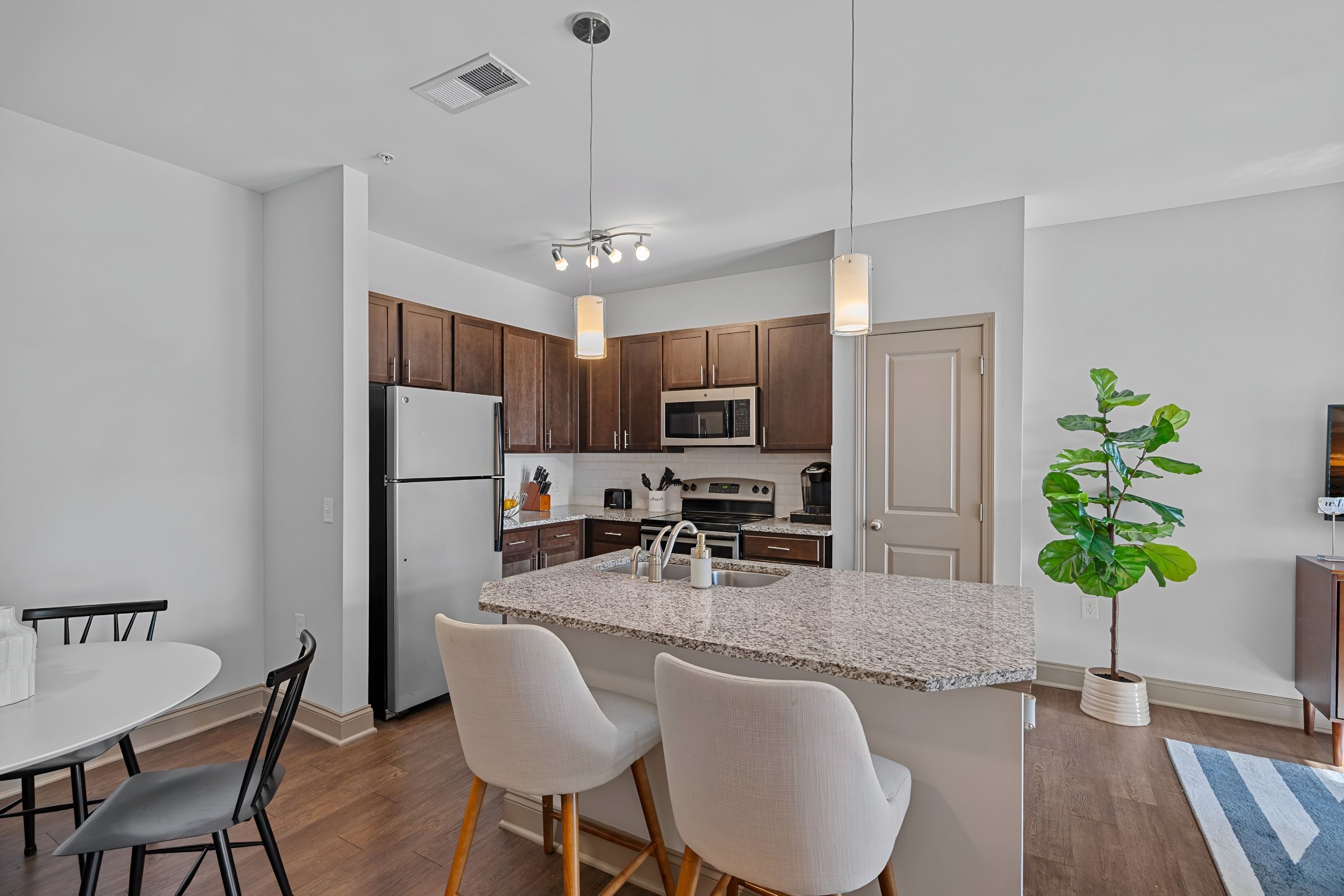 Modern kitchen with granite island, two white stools, stainless steel appliances, dark cabinets, and a potted plant in a bright open space.