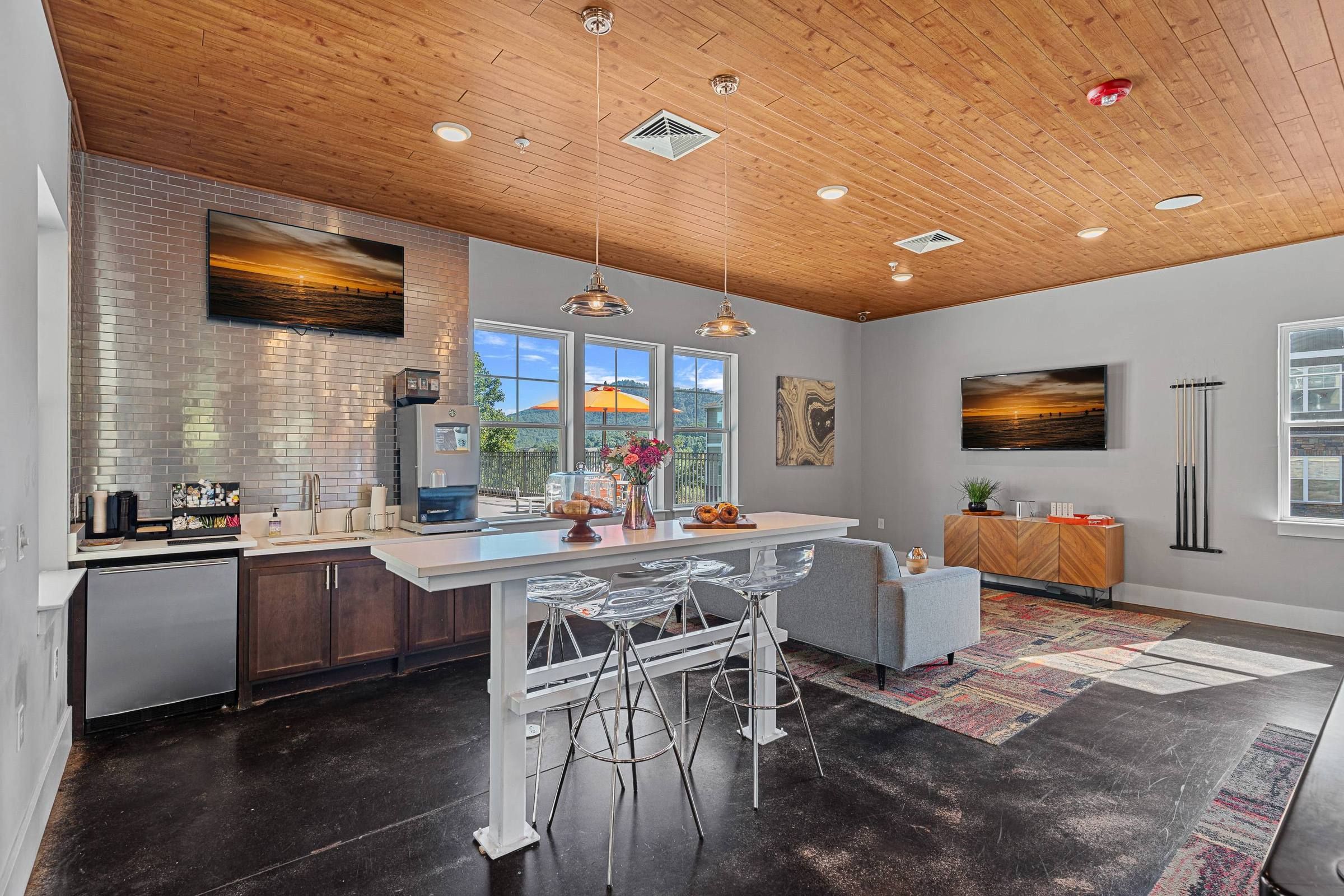 Modern lounge area with kitchen, high white table with clear stools, gray sofa, pendant lights, flat-screen TVs, and large windows.