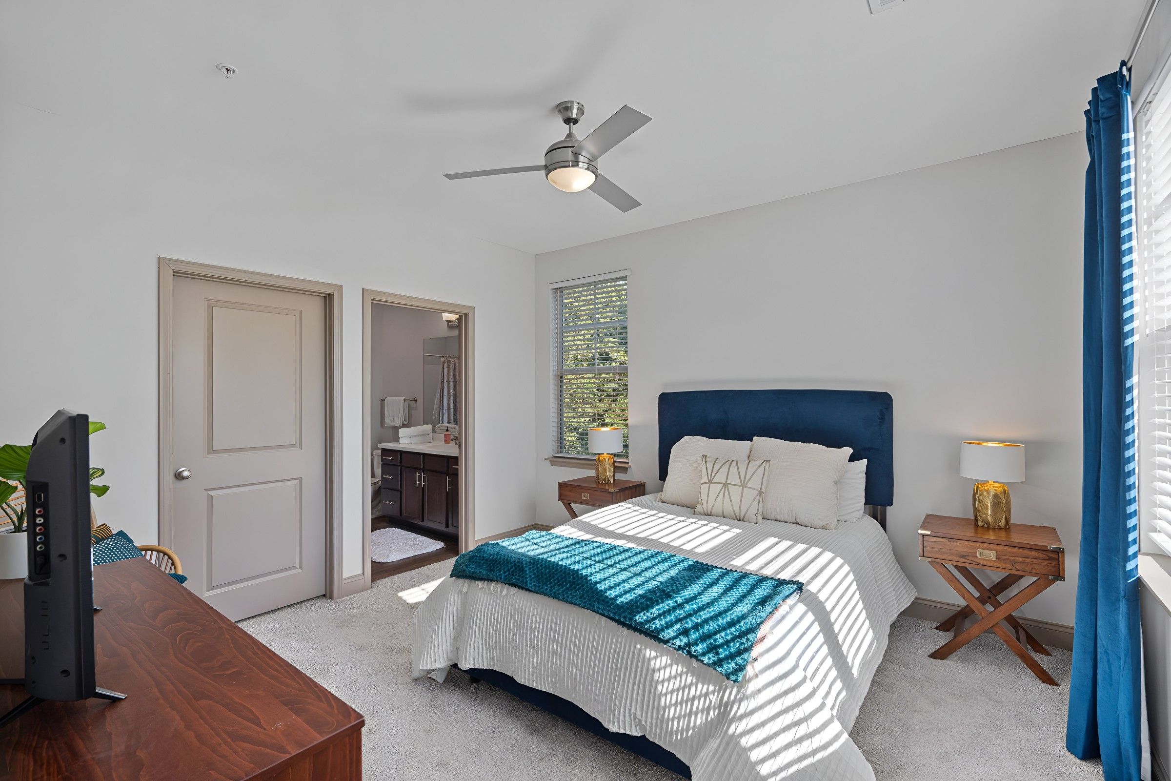 Bright bedroom with a blue upholstered bed, white bedding, teal throw, wooden nightstands, ceiling fan, and an adjoining bathroom.