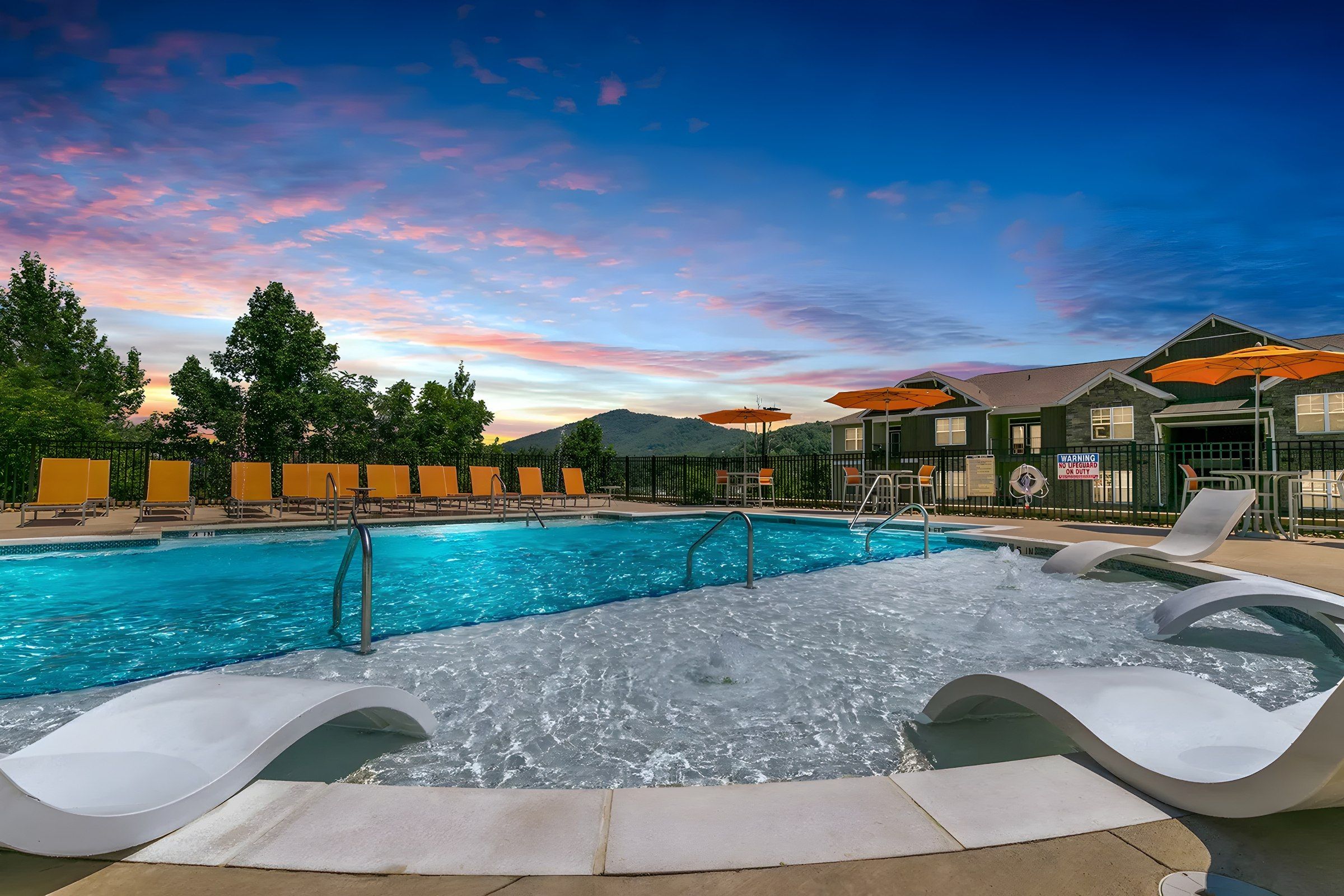 Outdoor swimming pool with lounge chairs and umbrellas at sunset, surrounded by trees and buildings with mountains in the background.