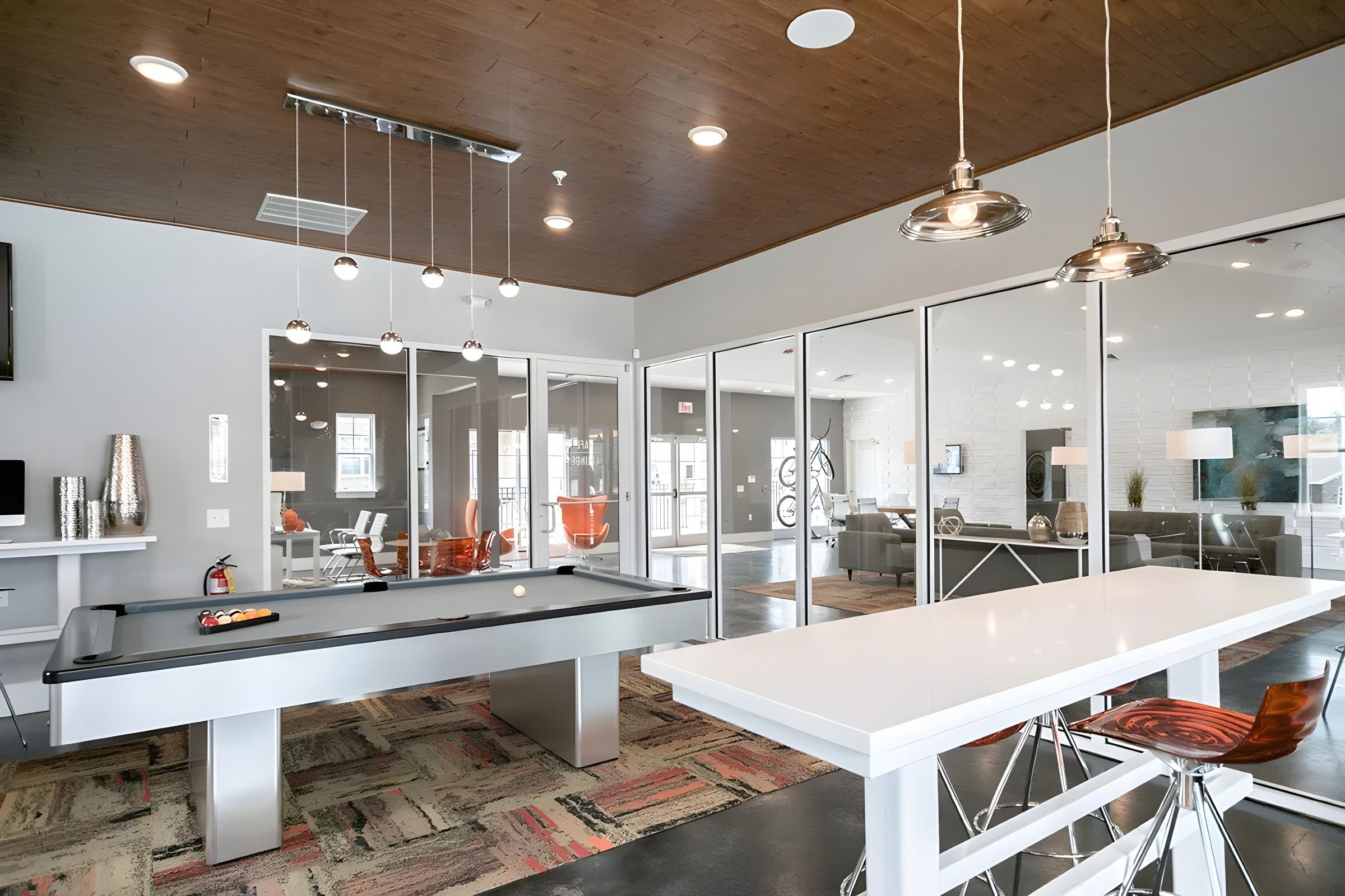 Modern game room with a pool table, white high table, wooden chair, pendant lights, and glass walls revealing adjacent seating areas.