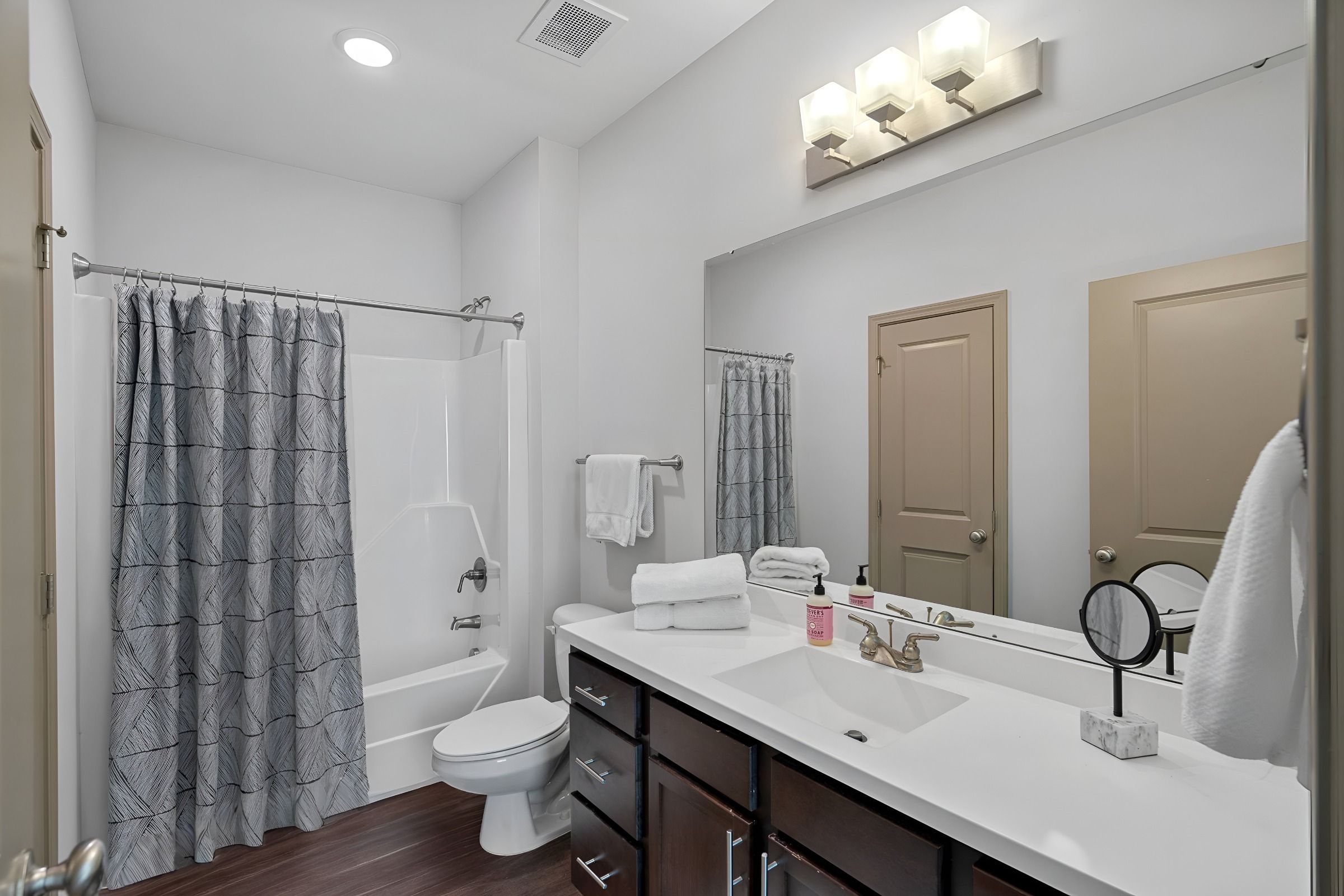 Modern bathroom with white countertop, dark cabinets, large mirror, towel racks, shower with patterned curtain, and wood-style flooring.